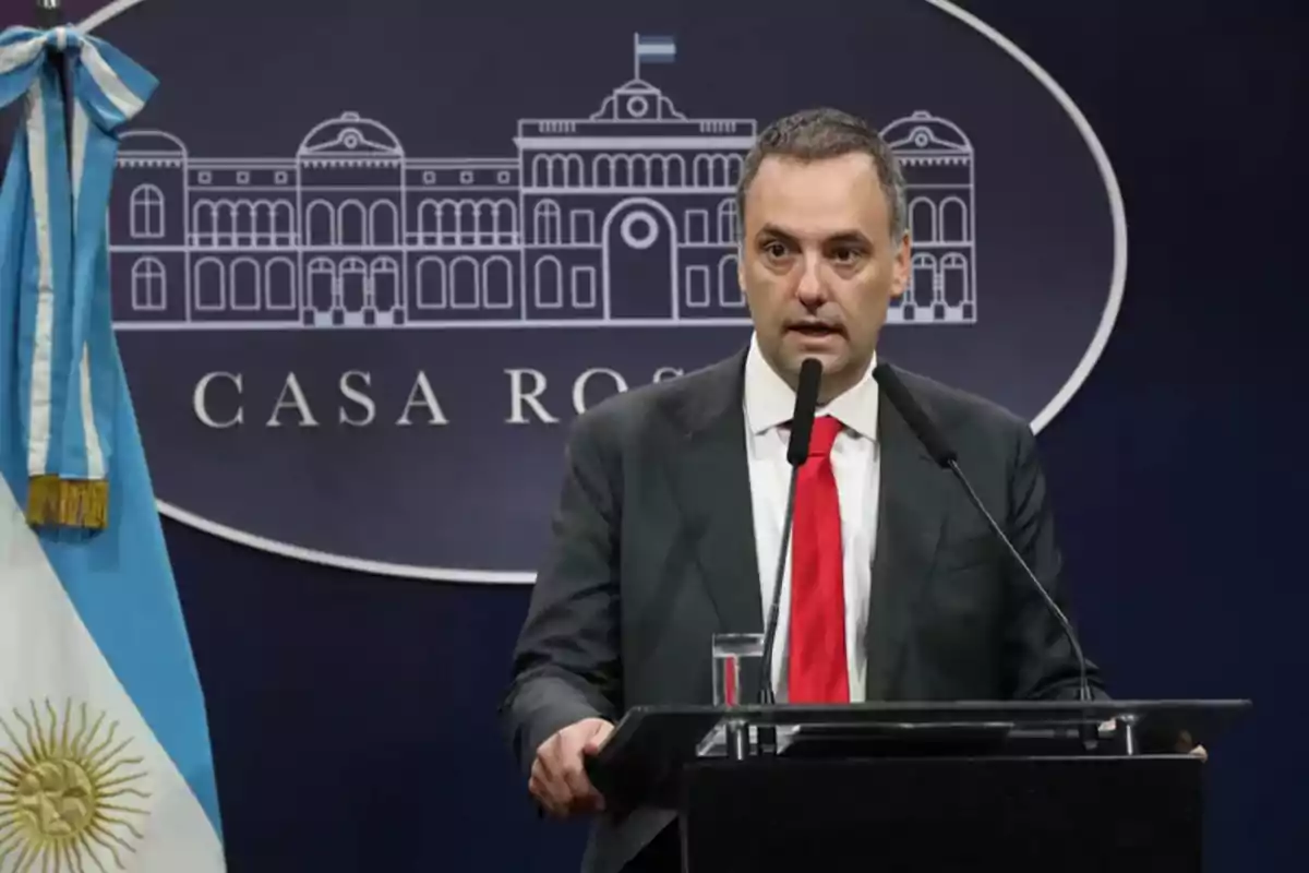 Un hombre de traje y corbata roja hablando en un podio con un micrófono, con una bandera argentina a su izquierda y un fondo que dice "Casa Rosada".