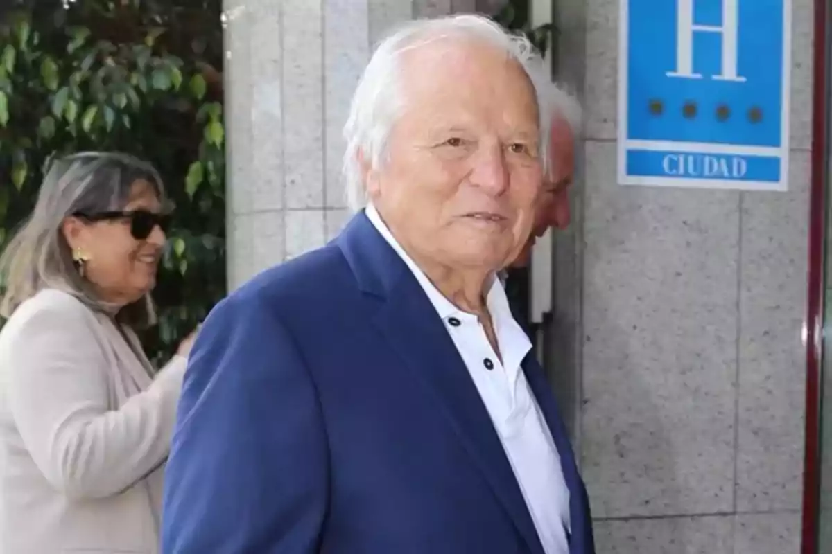 Manuel Benítez con cabello blanco y traje azul está de pie frente a un edificio con una mujer sonriendo en el fondo. Manuel Benítez con cabello blanco y traje azul está de pie frente a un edificio con una mujer sonriendo en el fondo.
