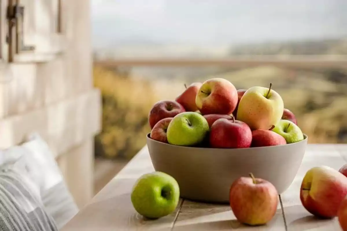 Un tazón lleno de manzanas de diferentes colores sobre una mesa de madera con un paisaje borroso al fondo.