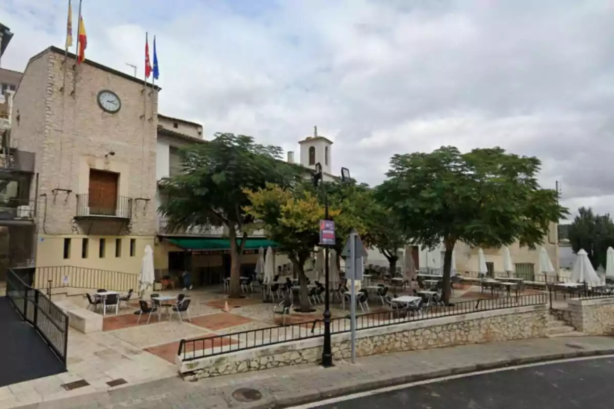 Una plaza con mesas y sillas al aire libre rodeada de árboles y un edificio de piedra con un reloj y varias banderas en la parte superior. Una plaza con mesas y sillas al aire libre rodeada de árboles y un edificio de piedra con un reloj y varias banderas en la parte superior.