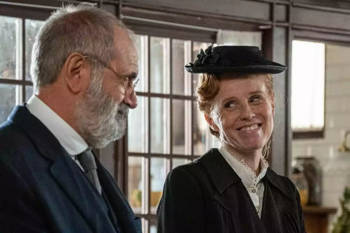 Joaquín Climent mayor con barba y María Castro con sombrero sonríen mientras están de pie en una habitación con ventanas grandes en La Promesa.