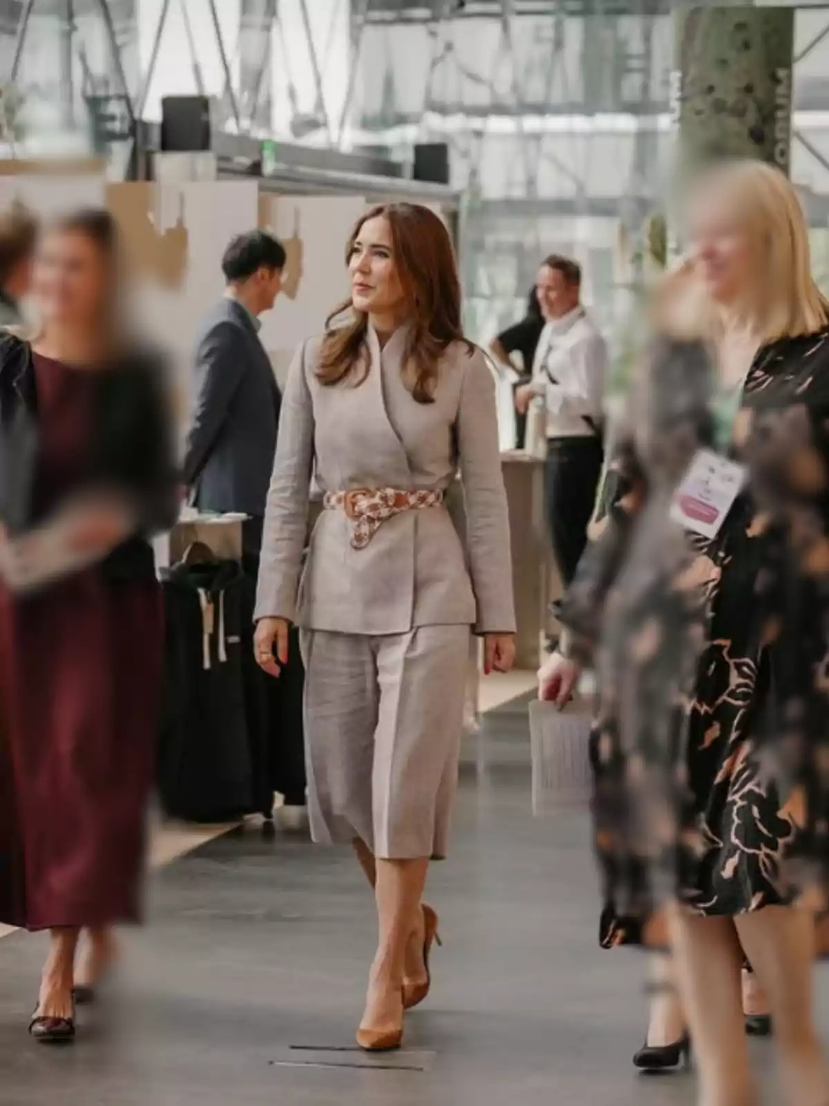 Brown-haired woman dressed in a beige outfit and brown heels walking in a modern indoor space surrounded by other people Brown-haired woman dressed in a beige outfit and brown heels walking in a modern indoor space surrounded by other people