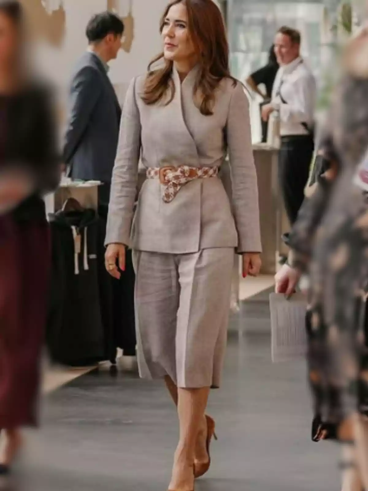 Mujer con cabello castaño claro usando un conjunto beige de chaqueta y falda midi, cinturón estampado y tacones nude, caminando en un espacio interior con otras personas alrededor. Mujer con cabello castaño claro usando un conjunto beige de chaqueta y falda midi, cinturón estampado y tacones nude, caminando en un espacio interior con otras personas alrededor.