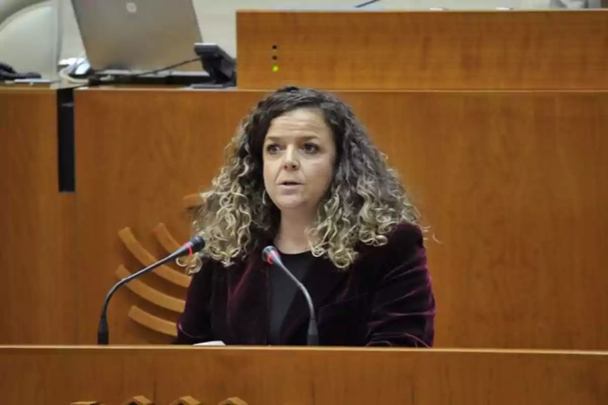 Una mujer con cabello rizado y rubio oscuro está hablando en un podio de madera con micrófonos frente a ella en un entorno formal. Una mujer con cabello rizado y rubio oscuro está hablando en un podio de madera con micrófonos frente a ella en un entorno formal.