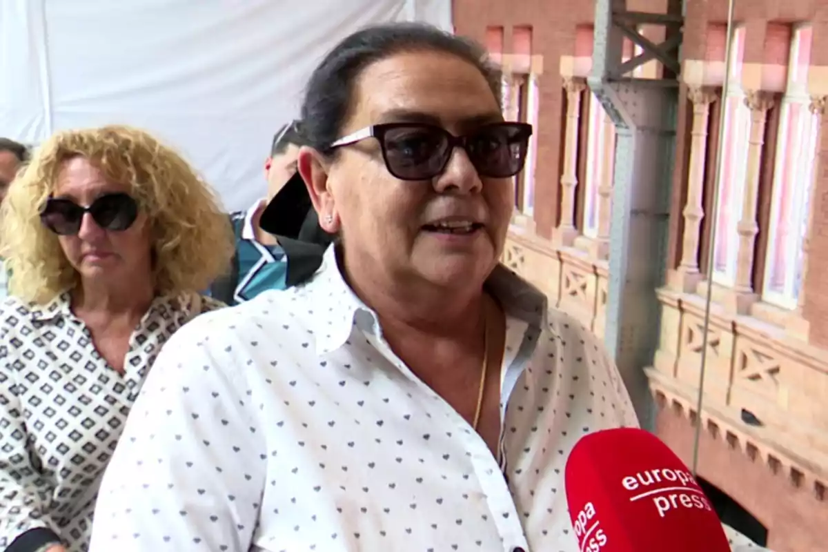 María del Monte con gafas de sol y camisa blanca con pequeños estampados siendo entrevistada por un micrófono rojo de Europa Press, con otra mujer al fondo y un edificio de ladrillo detrás. María del Monte con gafas de sol y camisa blanca con pequeños estampados siendo entrevistada por un micrófono rojo de Europa Press, con otra mujer al fondo y un edificio de ladrillo detrás.