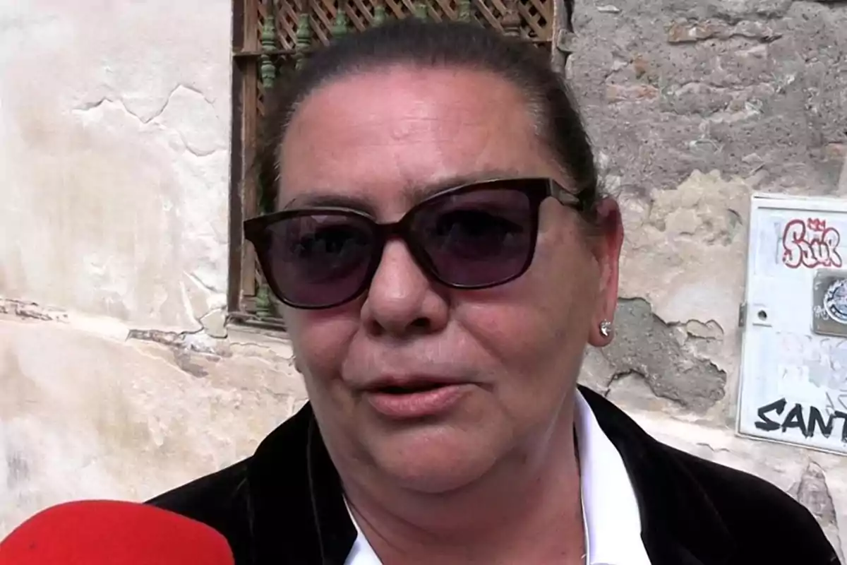 María del Monte con gafas de sol y chaqueta oscura habla frente a un micrófono rojo con una pared de fondo. María del Monte con gafas de sol y chaqueta oscura habla frente a un micrófono rojo con una pared de fondo.