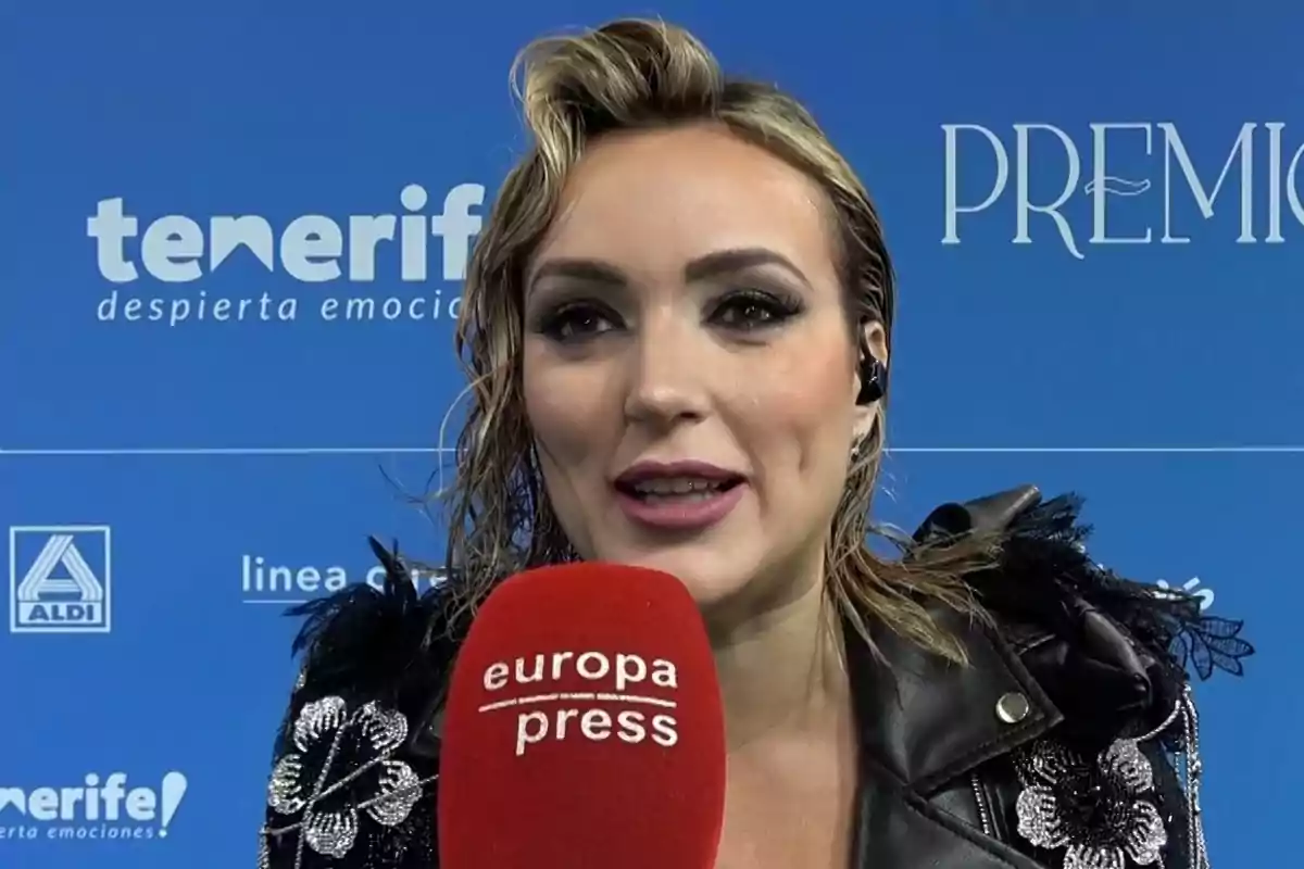 Marta Riesco con chaqueta negra decorada sostiene un micrófono rojo de Europa Press frente a un fondo azul con logotipos de Tenerife y Aldi. Marta Riesco con chaqueta negra decorada sostiene un micrófono rojo de Europa Press frente a un fondo azul con logotipos de Tenerife y Aldi.