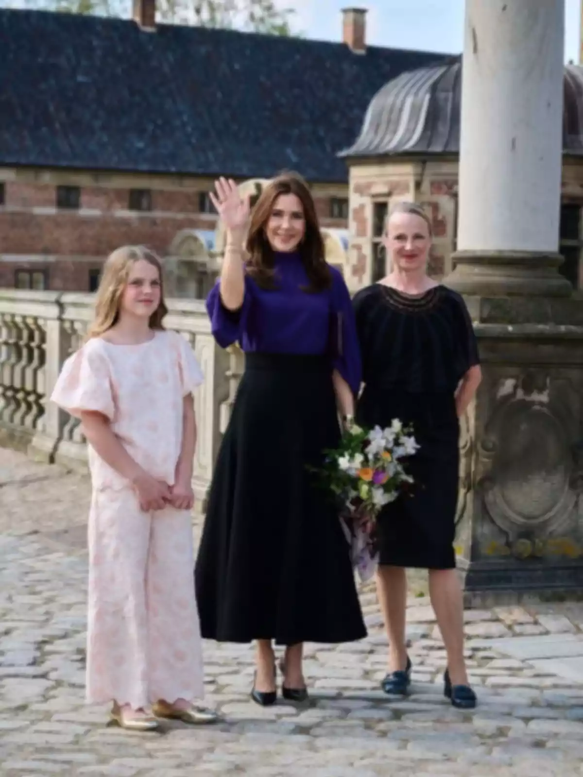 Three people pose outdoors in a historical setting, one of them waves while holding a bouquet of flowers. Three people pose outdoors in a historical setting, one of them waves while holding a bouquet of flowers.