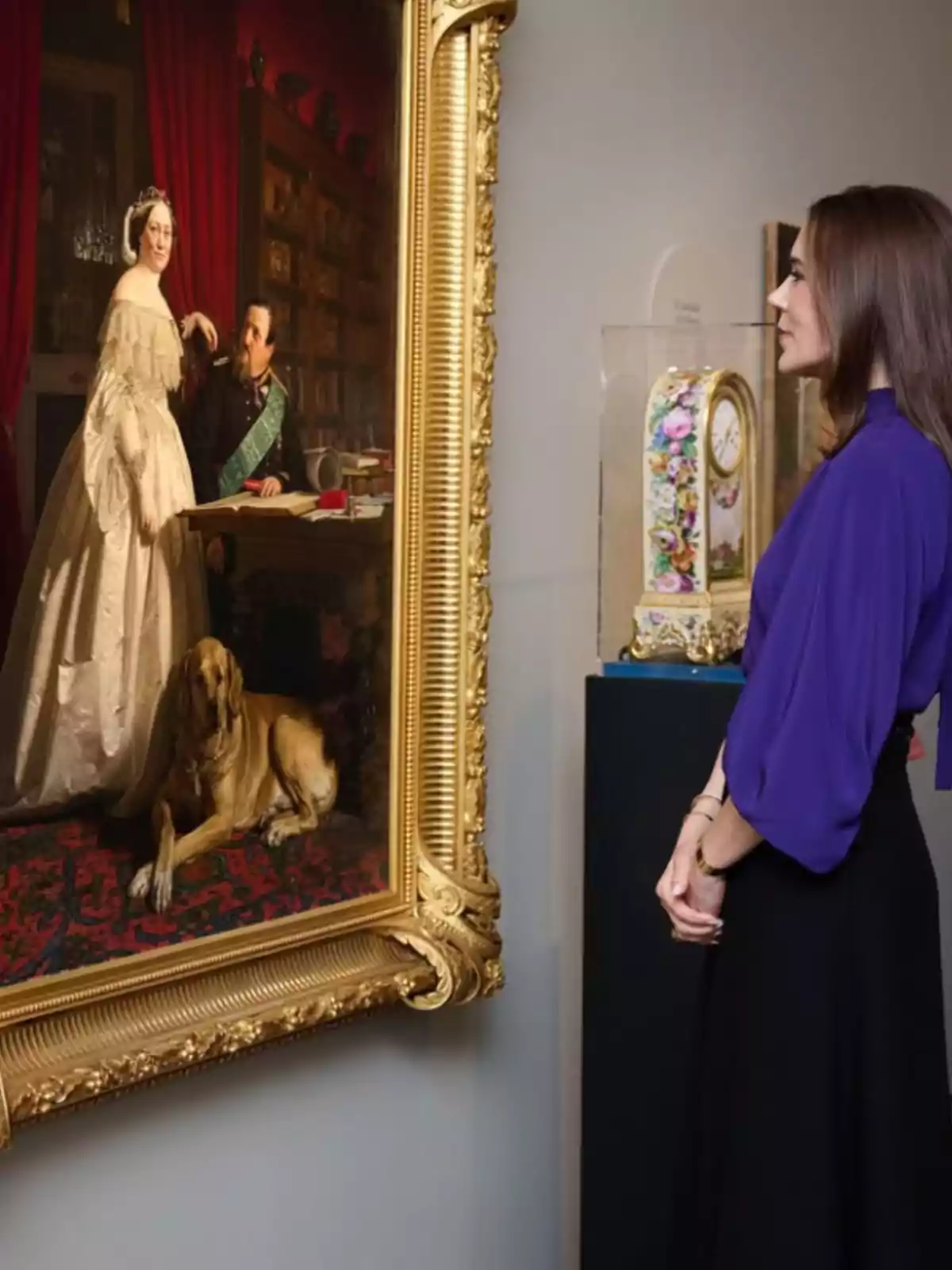 A woman closely observes a large framed painting depicting an elegant couple and a dog in a decorated room. A woman closely observes a large framed painting depicting an elegant couple and a dog in a decorated room.