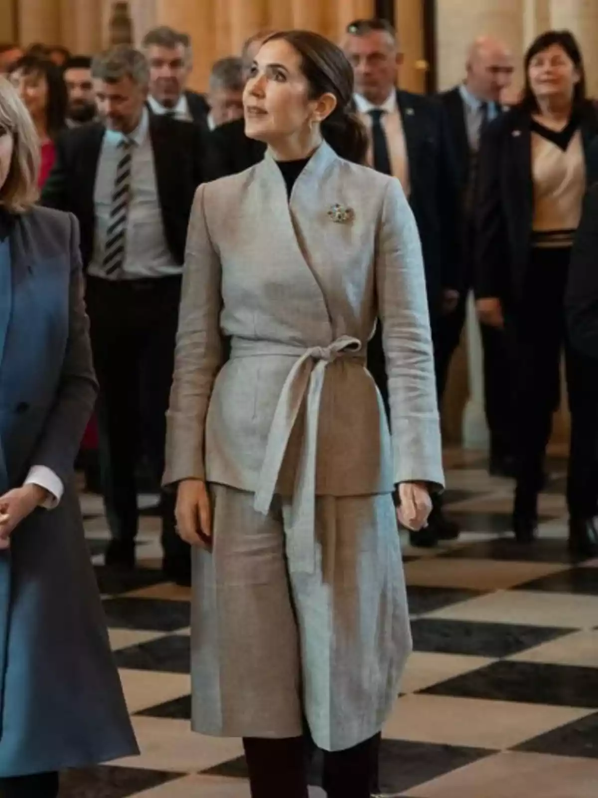 Mujer vestida con un conjunto gris claro y broche, de pie en un lugar elegante con piso de cuadros, rodeada de varias personas en traje formal Mujer vestida con un conjunto gris claro y broche, de pie en un lugar elegante con piso de cuadros, rodeada de varias personas en traje formal