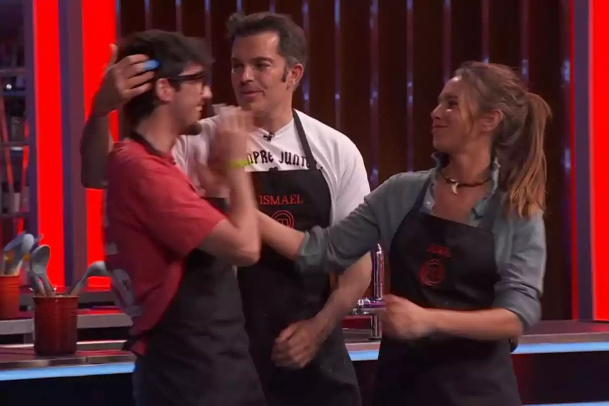 Emilio, Ismael y Ana con delantales de cocina interactúan en un set de competencia culinaria, una mujer sonríe mientras toca el rostro de un hombre con gafas y otro hombre los observa. Captura de 'MasterChef' Emilio, Ismael y Ana con delantales de cocina interactúan en un set de competencia culinaria, una mujer sonríe mientras toca el rostro de un hombre con gafas y otro hombre los observa. Captura de 'MasterChef'