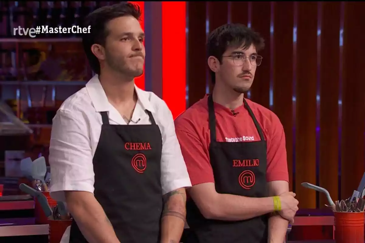 Dos concursantes de 'MasterChef' con delantales negros y nombres bordados en rojo están de pie en la cocina del programa con expresión seria. Dos concursantes de 'MasterChef' con delantales negros y nombres bordados en rojo están de pie en la cocina del programa con expresión seria.