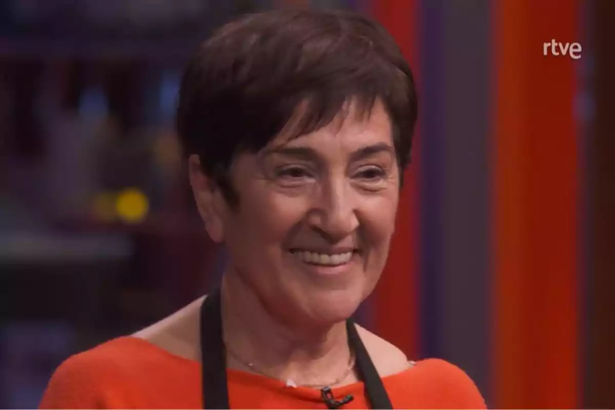 Ana María sonriente con cabello corto y oscuro lleva un micrófono de solapa y un delantal negro sobre una prenda roja, con un fondo desenfocado y el logo de RTVE en la esquina superior derecha. Captura de 'MasterChef' Ana María sonriente con cabello corto y oscuro lleva un micrófono de solapa y un delantal negro sobre una prenda roja, con un fondo desenfocado y el logo de RTVE en la esquina superior derecha. Captura de 'MasterChef'