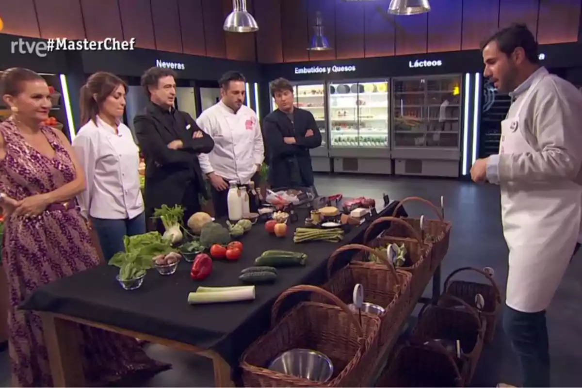 Un grupo de personas en un set de cocina de un programa de televisión observan una mesa con ingredientes frescos y cestas de mimbre, al fondo se ven refrigeradores con productos lácteos y embutidos. Captura de 'MasterChef' Un grupo de personas en un set de cocina de un programa de televisión observan una mesa con ingredientes frescos y cestas de mimbre, al fondo se ven refrigeradores con productos lácteos y embutidos. Captura de 'MasterChef'