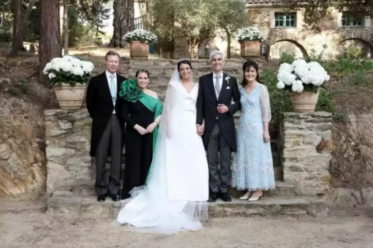 A group of elegantly dressed people posing for a photo in an outdoor setting with stone stairs and pots with white flowers. A group of elegantly dressed people posing for a photo in an outdoor setting with stone stairs and pots with white flowers.