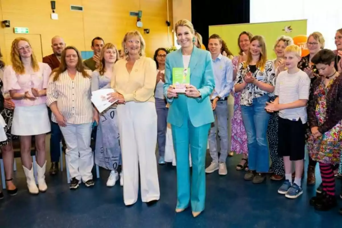 Un grupo de personas de pie en una sala, algunas aplaudiendo, mientras dos mujeres en el centro sostienen documentos y sonríen. Un grupo de personas de pie en una sala, algunas aplaudiendo, mientras dos mujeres en el centro sostienen documentos y sonríen.