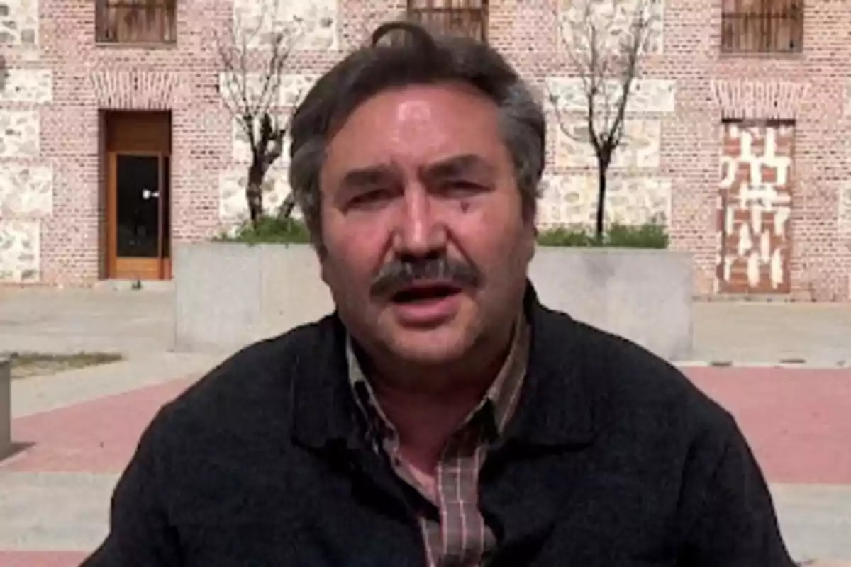 Un hombre con bigote está al aire libre frente a un edificio de ladrillo.