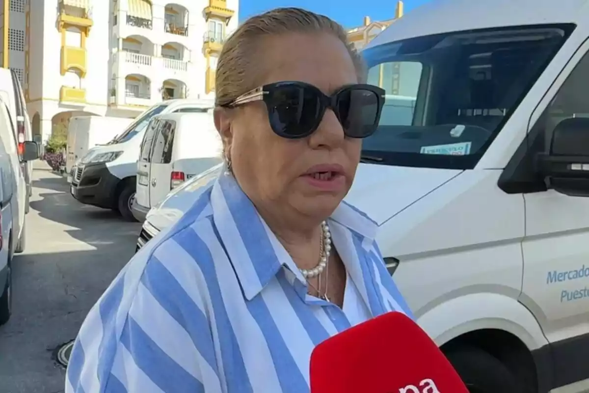 Mayte Zaldívar con gafas de sol y camisa a rayas es entrevistada en un estacionamiento con varias furgonetas y un edificio al fondo.