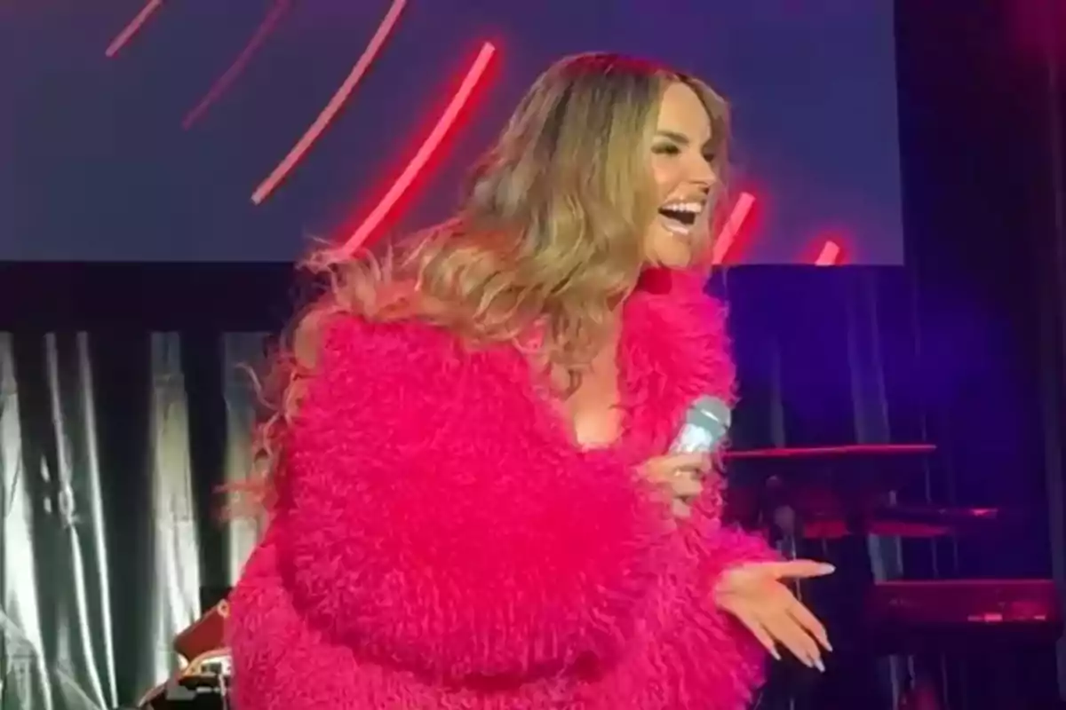 Melody con abrigo rosa brillante sonríe mientras sostiene un micrófono en un escenario iluminado Melody con abrigo rosa brillante sonríe mientras sostiene un micrófono en un escenario iluminado