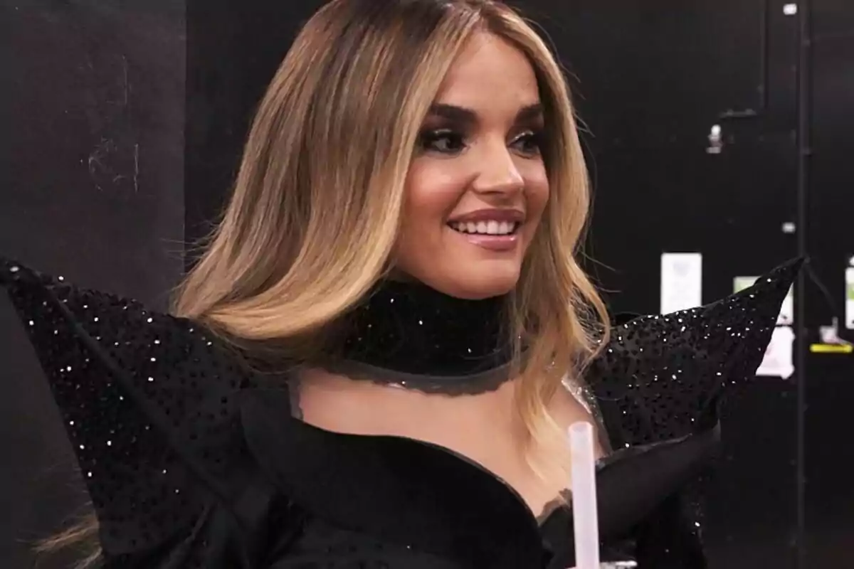 Melody con cabello largo y rubio sonriendo, vistiendo un traje negro con detalles brillantes y hombreras puntiagudas. Melody con cabello largo y rubio sonriendo, vistiendo un traje negro con detalles brillantes y hombreras puntiagudas.
