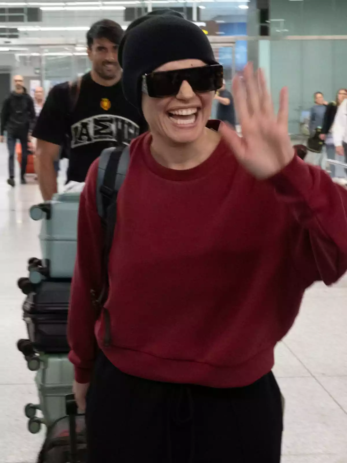 Melody sonriente con gafas de sol y gorro negro saluda mientras camina por un aeropuerto, con varias maletas detrás. Melody sonriente con gafas de sol y gorro negro saluda mientras camina por un aeropuerto, con varias maletas detrás.