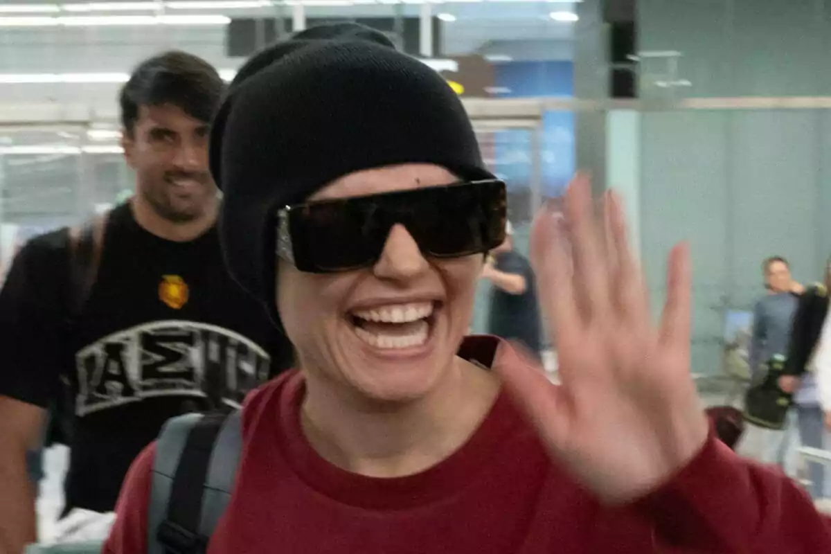 Melody sonriente con gafas de sol y gorro negro saluda mientras camina en su llegada al aeropuerto. Melody sonriente con gafas de sol y gorro negro saluda mientras camina en su llegada al aeropuerto.