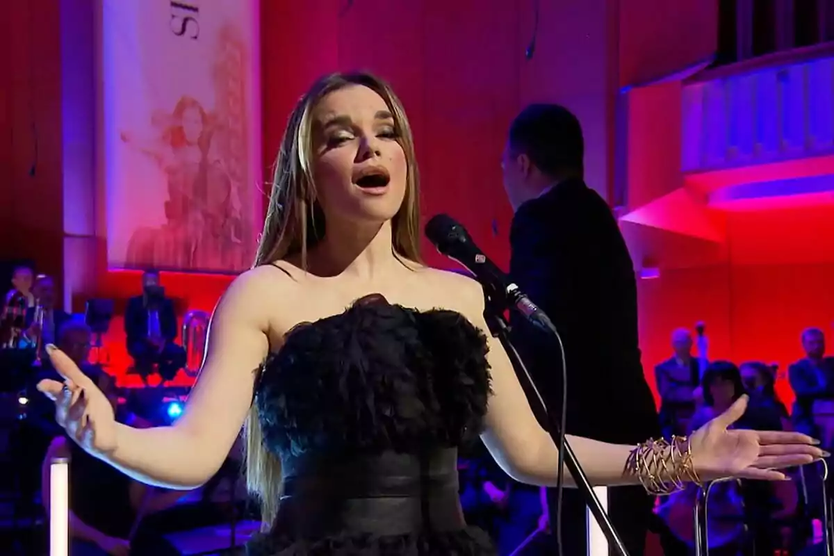 Melody canta apasionadamente en un escenario iluminado con luces de colores, mientras el director de Orquesta y Coro de RTVE se encuentra al fondo. Melody canta apasionadamente en un escenario iluminado con luces de colores, mientras el director de Orquesta y Coro de RTVE se encuentra al fondo.