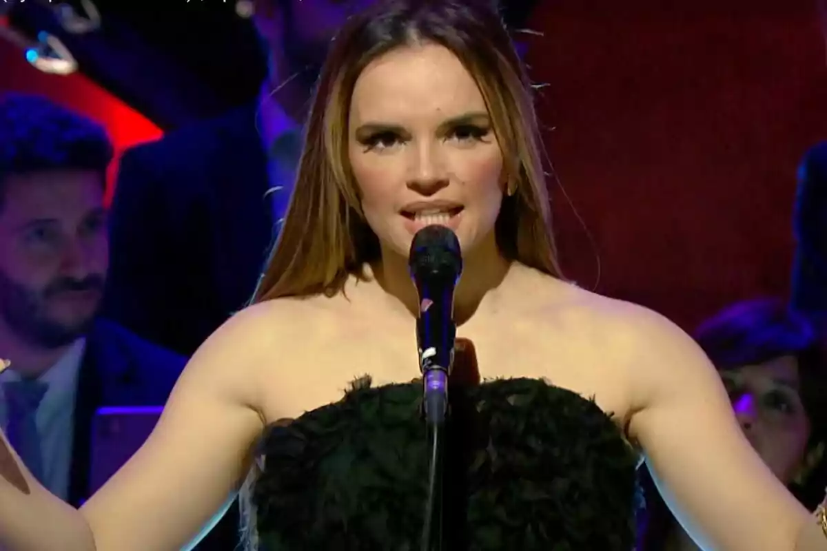 Melody canta en un escenario con un micrófono acompañada de la Orquesta Sinfónica de RTVE, vestida de negro, mientras el fondo está iluminado con luces de colores. Melody canta en un escenario con un micrófono acompañada de la Orquesta Sinfónica de RTVE, vestida de negro, mientras el fondo está iluminado con luces de colores.