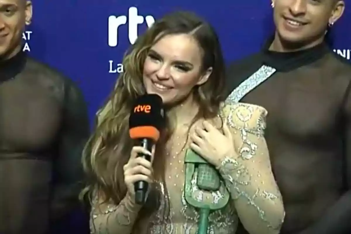 Melody sonriente sostiene un micrófono mientras está acompañada por dos personas en un evento con fondo azul en la rueda de prensa tras el Benidorm Fest. Melody sonriente sostiene un micrófono mientras está acompañada por dos personas en un evento con fondo azul en la rueda de prensa tras el Benidorm Fest.