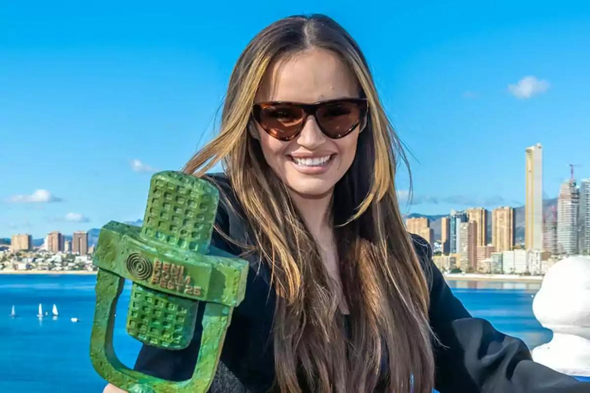 Melody con gafas de sol sonríe mientras sostiene un trofeo verde con la inscripción Melody con gafas de sol sonríe mientras sostiene un trofeo verde con la inscripción