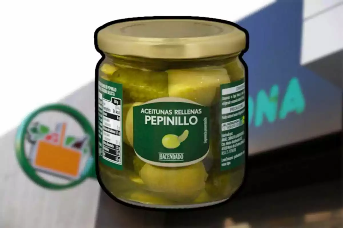 Un frasco de aceitunas rellenas de pepinillo de la marca Hacendado frente a un cartel borroso de supermercado. Un frasco de aceitunas rellenas de pepinillo de la marca Hacendado frente a un cartel borroso de supermercado.