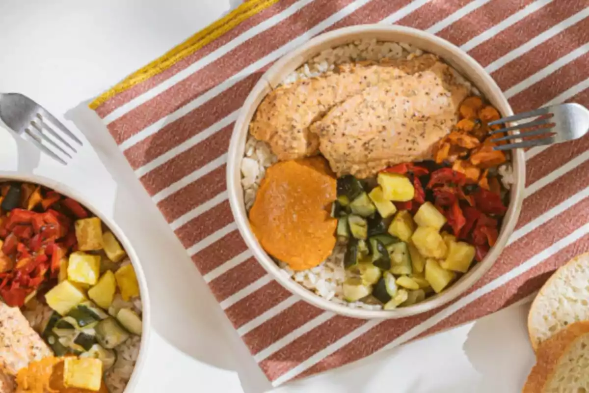 Dos tazones de comida con arroz, vegetales asados y una salsa, sobre un mantel a rayas rojas y blancas, con tenedores y pan al lado. Dos tazones de comida con arroz, vegetales asados y una salsa, sobre un mantel a rayas rojas y blancas, con tenedores y pan al lado.