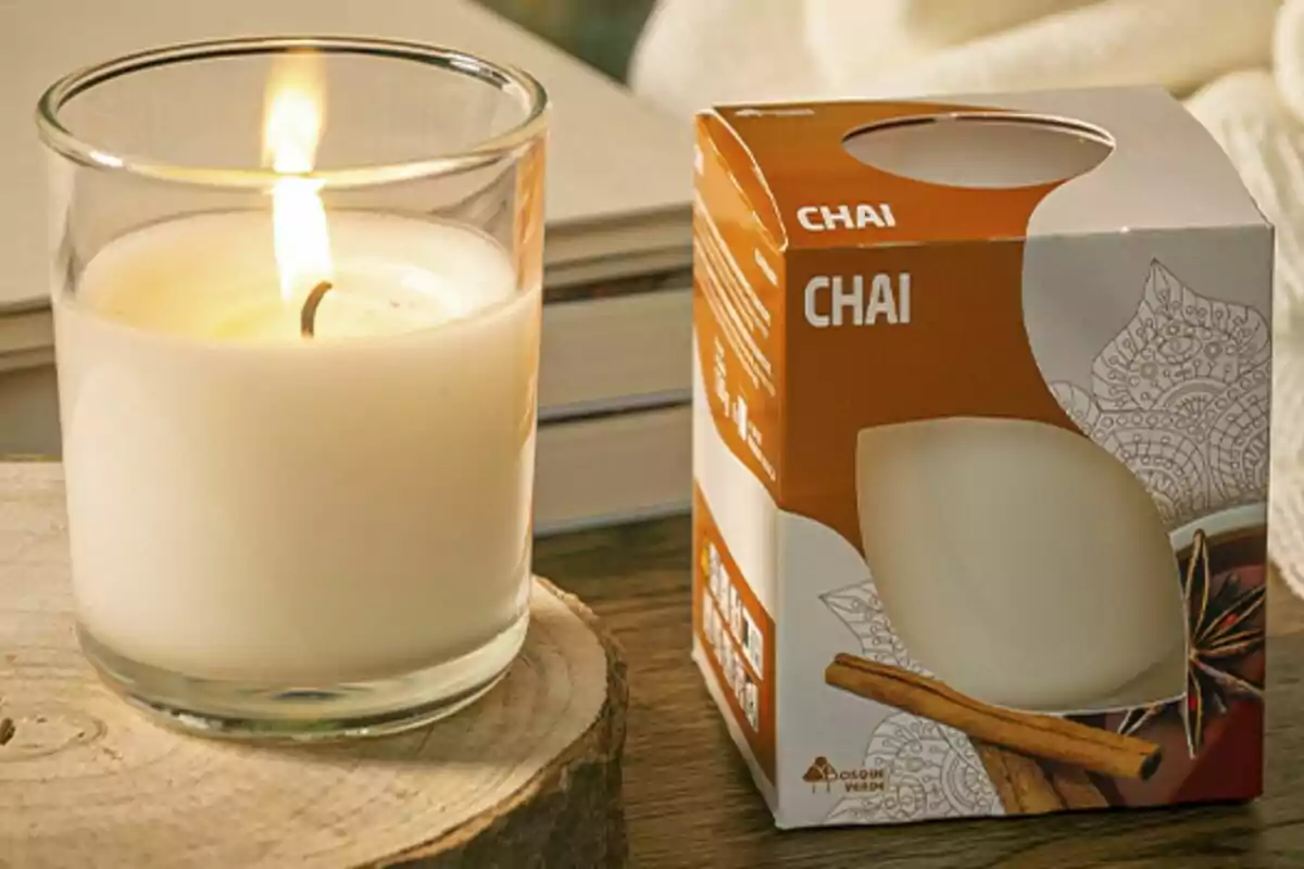 Vela aromática encendida en un vaso de vidrio junto a su caja de empaque con diseño de chai sobre una superficie de madera. Vela aromática encendida en un vaso de vidrio junto a su caja de empaque con diseño de chai sobre una superficie de madera.