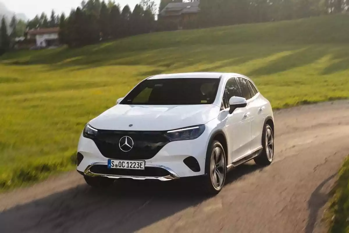 Un coche Mercedes-Benz blanco circulando por un camino en un entorno rural con césped verde y árboles al fondo.