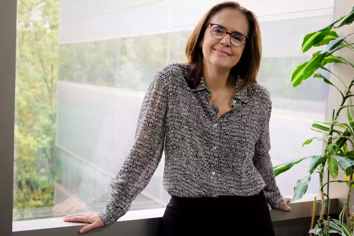 Una persona con gafas y camisa estampada está apoyada en el alféizar de una ventana, con plantas a su lado y un fondo de árboles visibles a través del cristal.