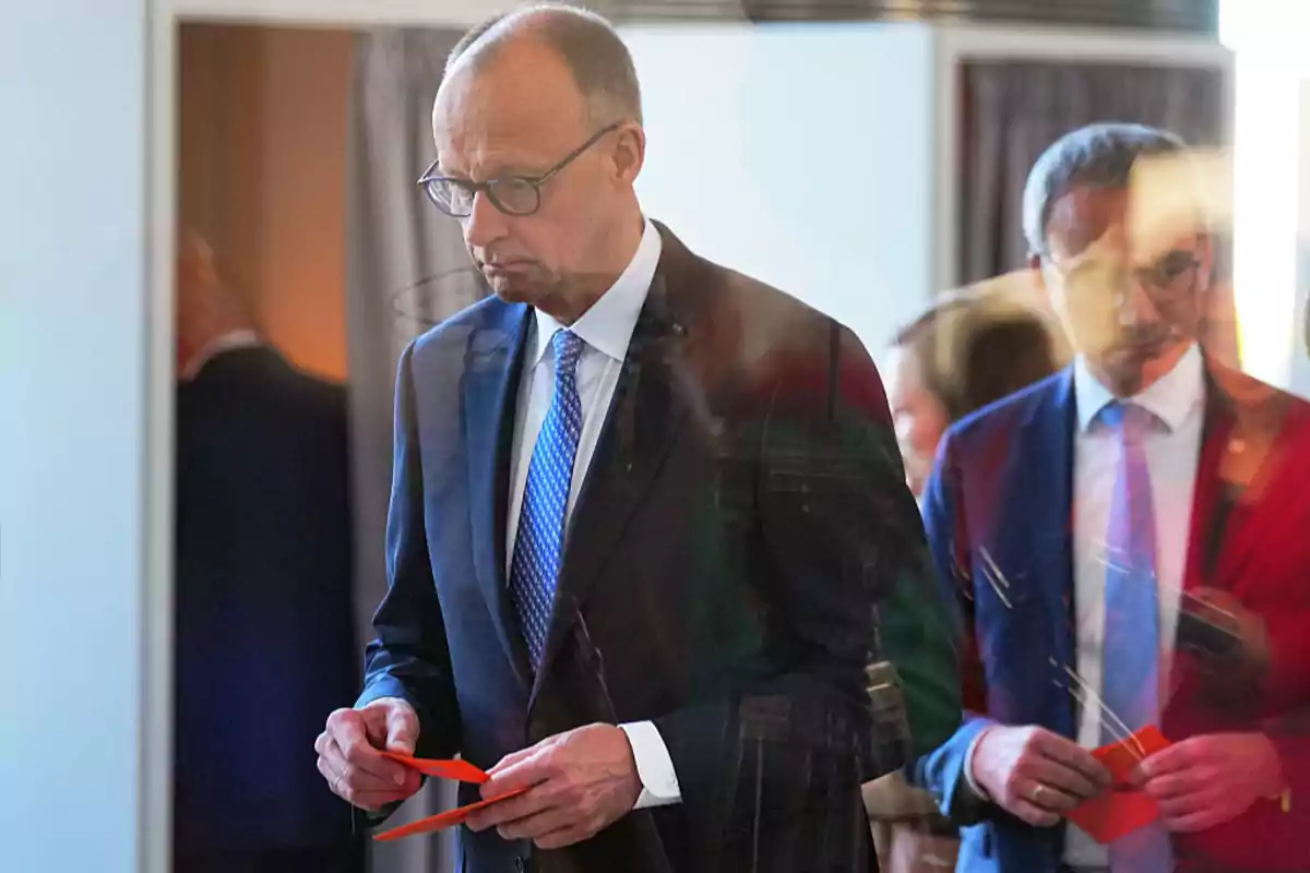 Un hombre con traje y corbata azul sostiene papeles rojos mientras está de pie en una sala con otras personas al fondo. Un hombre con traje y corbata azul sostiene papeles rojos mientras está de pie en una sala con otras personas al fondo.