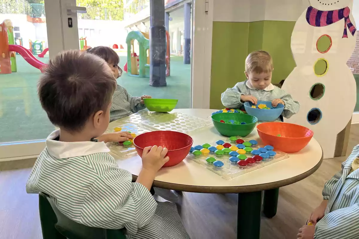 Niños pequeños jugando con fichas de colores en una mesa redonda dentro de un aula con un muñeco de nieve decorativo y un área de juegos visible a través de la ventana.