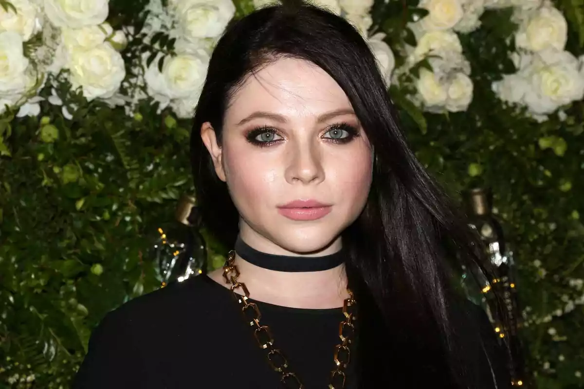 Una persona con cabello oscuro y ojos claros, usando un collar de cadena dorada, frente a un fondo de flores blancas y hojas verdes.