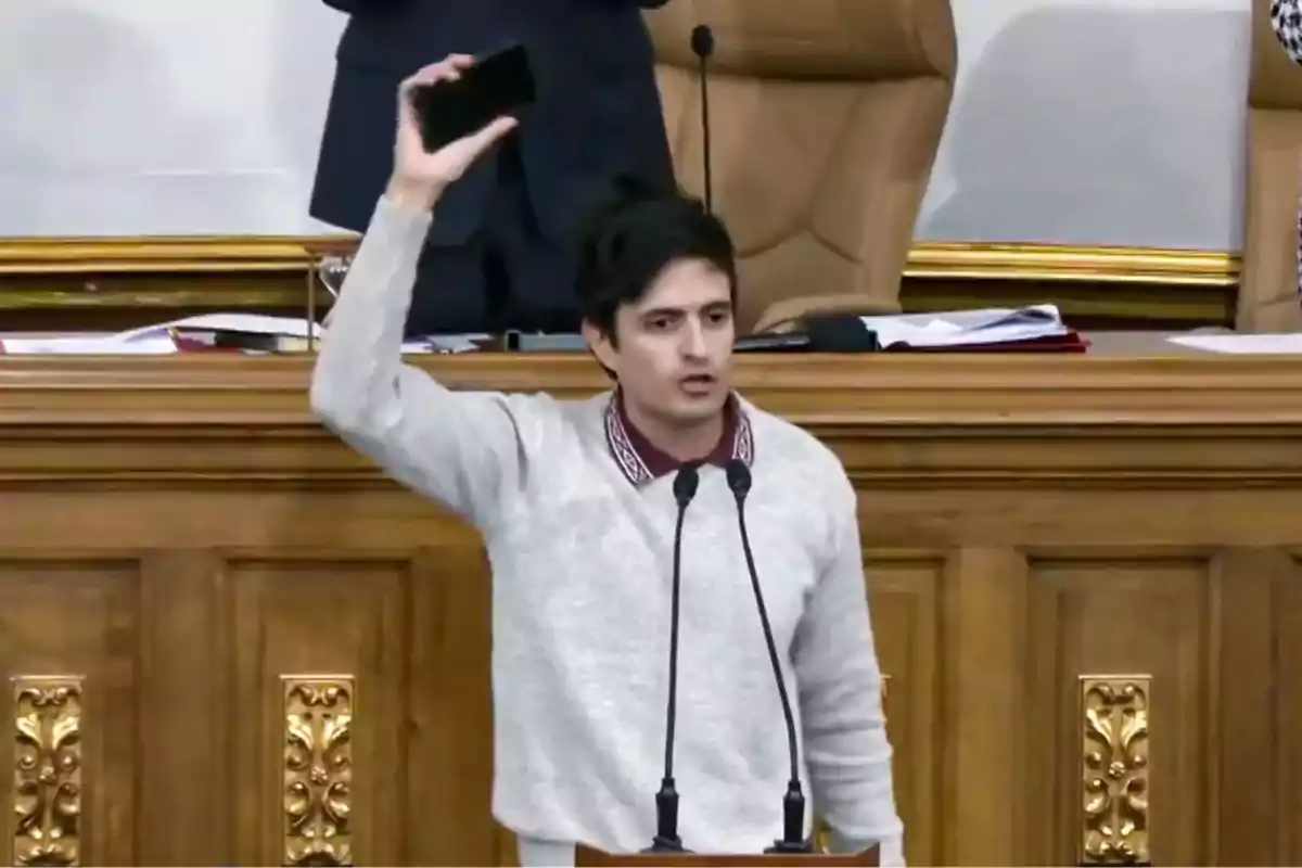 Un hombre de pie detrás de un podio de madera, levantando un teléfono móvil en su mano derecha, mientras habla frente a dos micrófonos en un entorno formal con sillas de respaldo alto y documentos sobre una mesa.