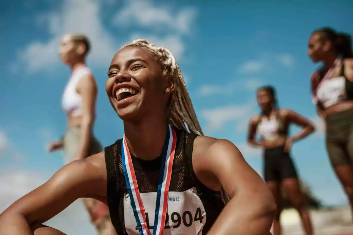 Una atleta sonriente con una medalla alrededor del cuello, sentada y rodeada de otras competidoras en un día soleado.