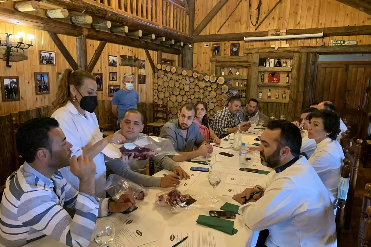 Un grupo de personas se encuentra en un restaurante de madera, sentados alrededor de una mesa larga, mientras una mujer con mascarilla negra les sirve comida.