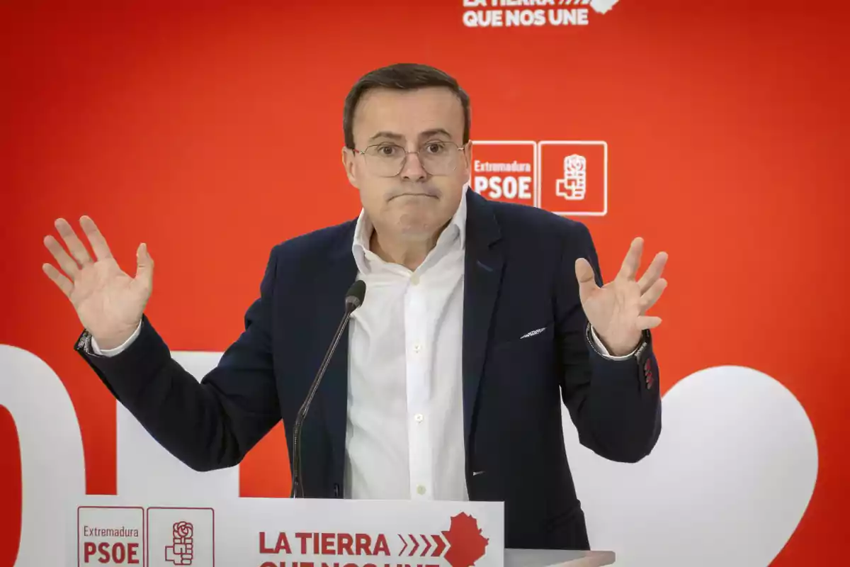 Un hombre con gafas y traje oscuro gesticula con las manos levantadas frente a un micrófono y un fondo rojo con logotipos del PSOE.