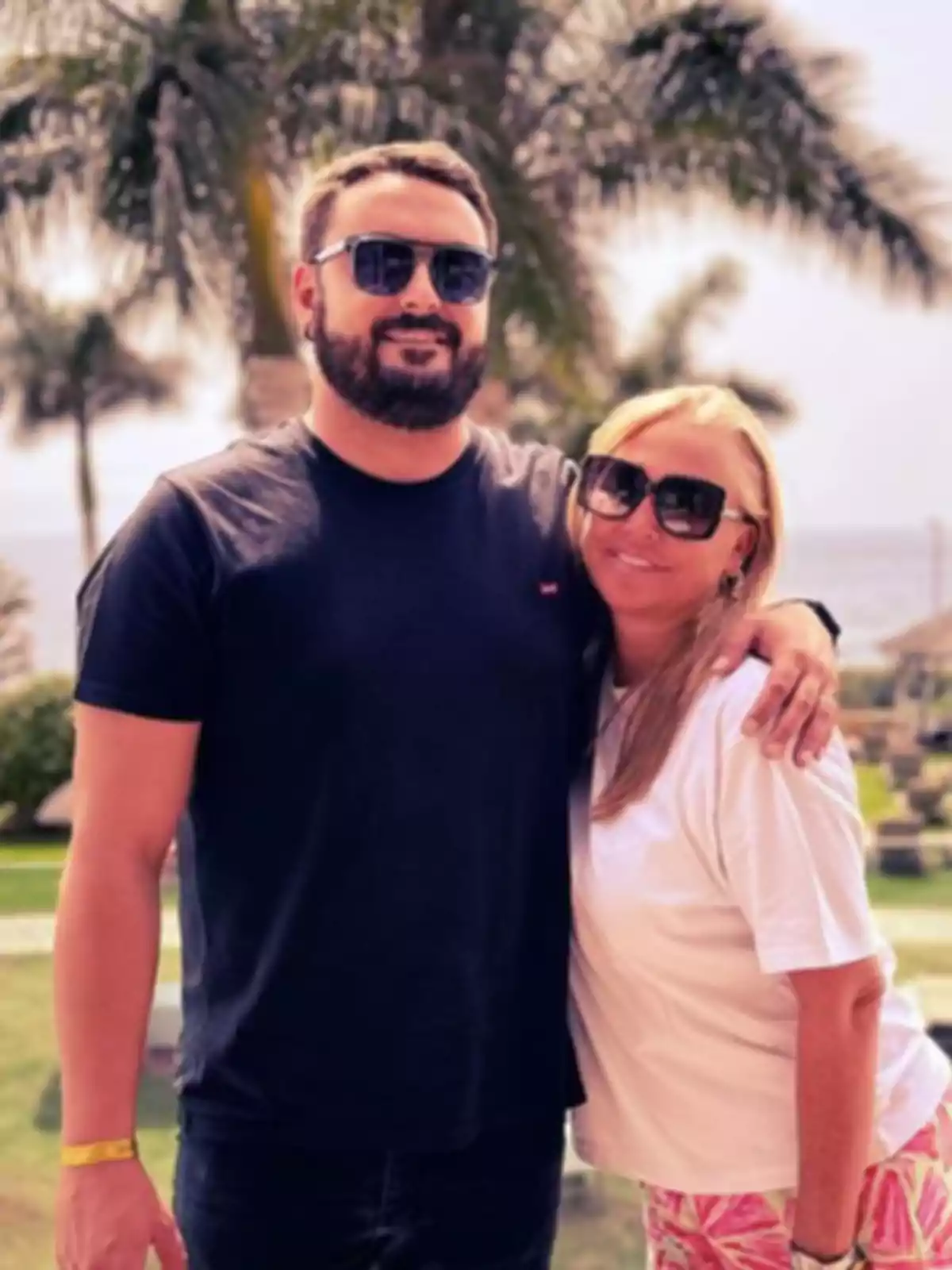 Miguel Marcos y Belén Esteban sonrientes con gafas de sol posan frente a un paisaje tropical con palmeras.