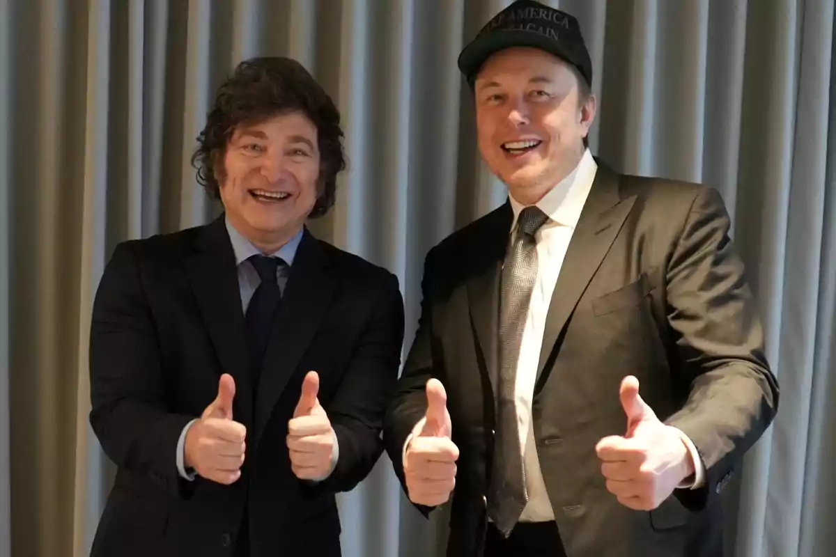 Dos hombres sonrientes con trajes oscuros levantan los pulgares frente a una cortina gris.