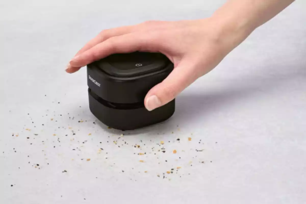 Una mano usando un pequeño aspirador de mesa negro para recoger migas sobre una superficie gris. Una mano usando un pequeño aspirador de mesa negro para recoger migas sobre una superficie gris.