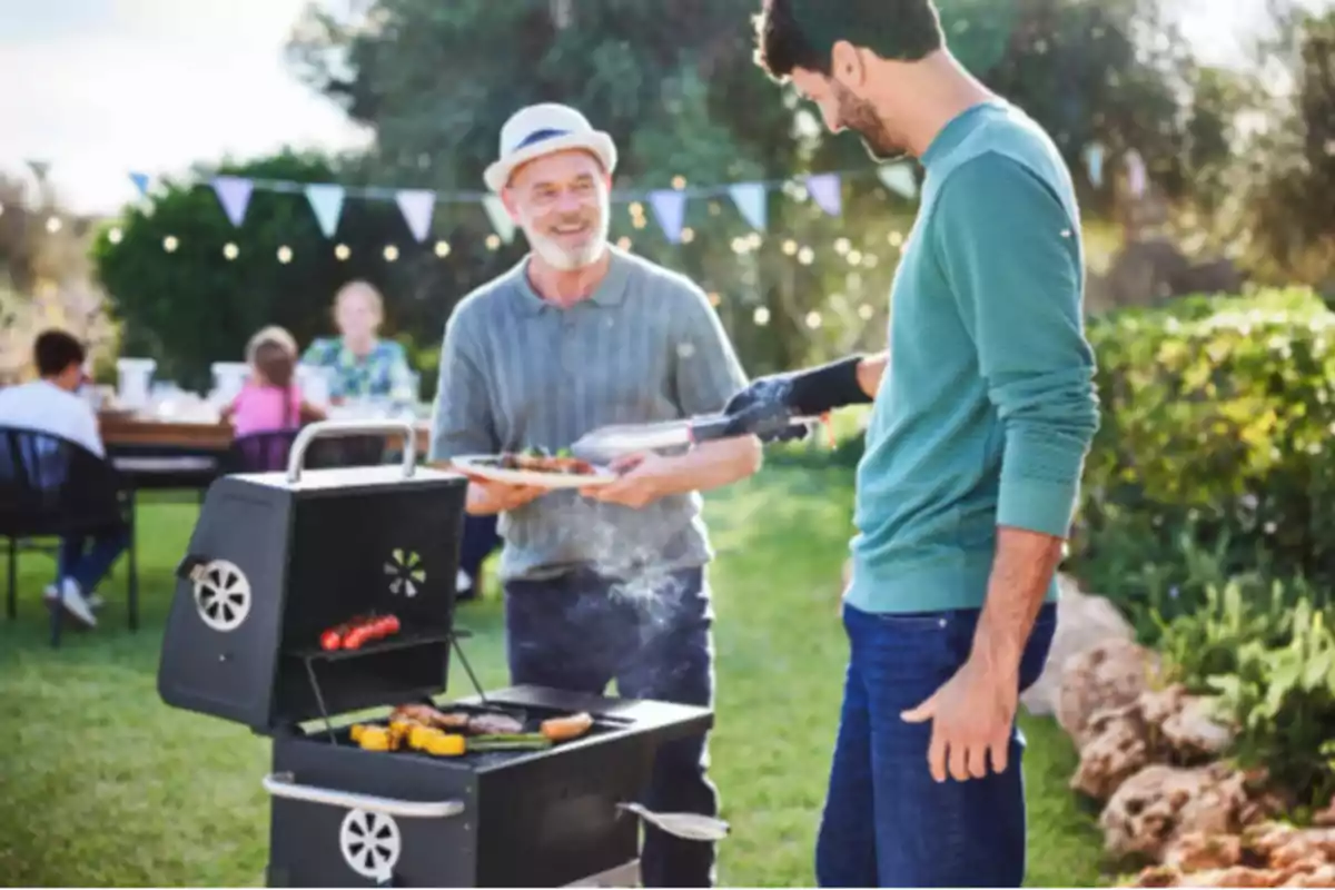 Dos personas cocinando en una parrilla al aire libre mientras otras personas están sentadas en una mesa en el fondo. Dos personas cocinando en una parrilla al aire libre mientras otras personas están sentadas en una mesa en el fondo.