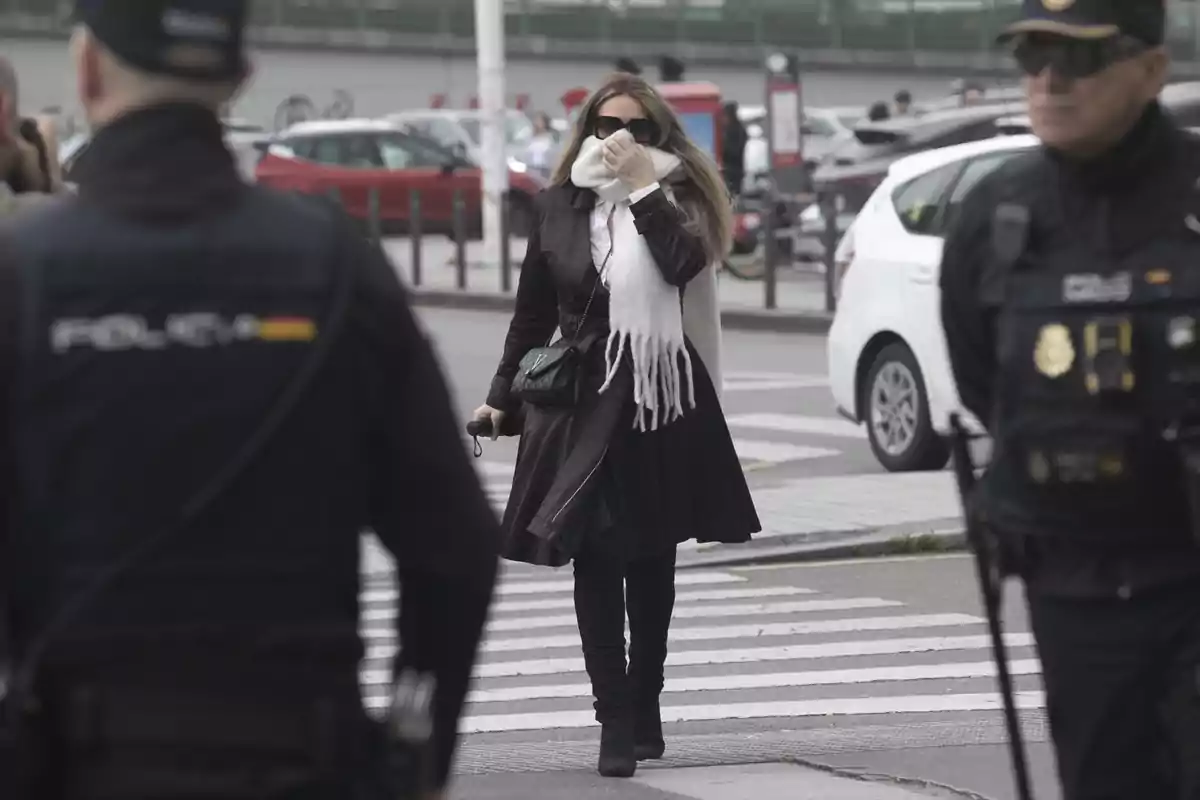 Una mujer con abrigo negro y bufanda blanca camina por un paso de peatones mientras dos policías están de pie cerca. Una mujer con abrigo negro y bufanda blanca camina por un paso de peatones mientras dos policías están de pie cerca.
