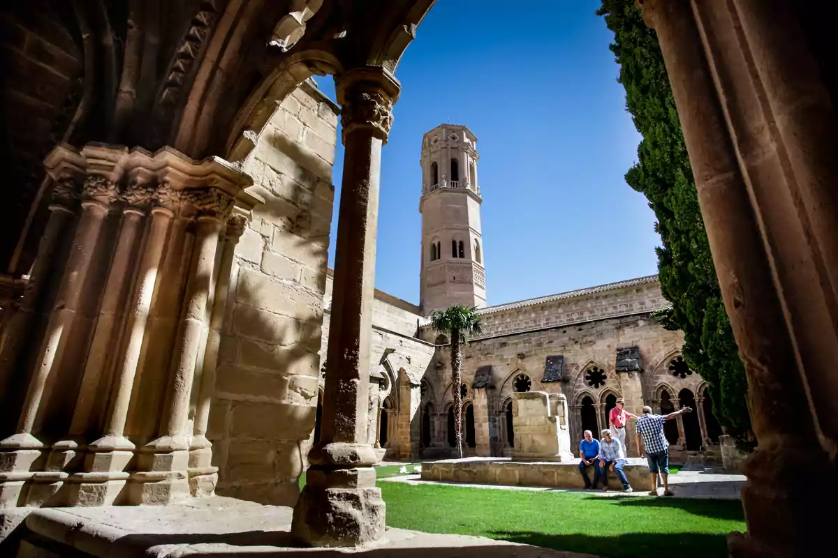 Un grupo de personas disfruta de un día soleado en un antiguo claustro con columnas y un campanario al fondo. Un grupo de personas disfruta de un día soleado en un antiguo claustro con columnas y un campanario al fondo.