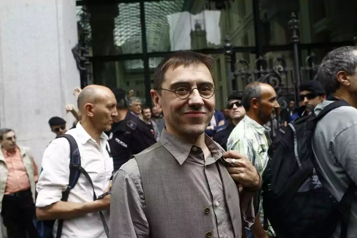 A man with glasses and a vest smiles at the camera while surrounded by people on a busy street.