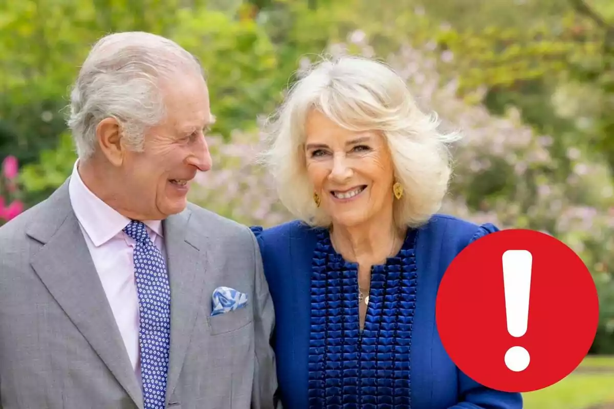 Carlos III y Camila Parker sonriente, el hombre con traje gris y corbata azul, la mujer con vestido azul, en un entorno al aire libre con vegetación de fondo y un icono de exclamación roja en la esquina inferior derecha.